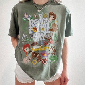 May include: A sage green short-sleeve t-shirt featuring a graphic print with the text "REACH FOR THE SKY" and various cartoon characters and icons from the Toy Story franchise, including Woody, Buzz Lightyear, Jessie, Rex, and the Pizza Planet truck.