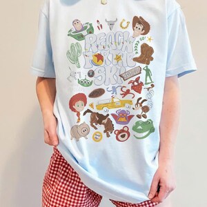 May include: A light blue t-shirt featuring a colourful graphic print of various animated characters and objects, including a cowboy, space ranger, dinosaur, and pig. The text "REACH FOR THE SKY" is printed in the centre in a wavy, light-coloured font.