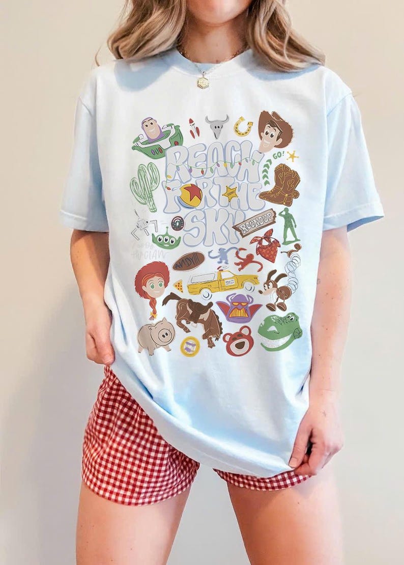 May include: A light blue t-shirt featuring a colourful graphic print of various animated characters and objects, including a cowboy, space ranger, dinosaur, and pig. The text "REACH FOR THE SKY" is printed in the centre in a wavy, light-coloured font.