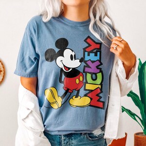 May include: Blue t-shirt featuring a colorful graphic of Mickey Mouse with the word "MICKEY" in vertical text. Mickey Mouse is depicted in his classic outfit with red shorts, yellow shoes, and black ears. The shirt has short sleeves.