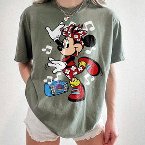 Pu&ograve; includere: T-shirt verde oliva con una grafica di Minnie Mouse che balla con note musicali e un boombox. Minnie Mouse indossa un vestito floreale rosso e bianco, scarpe rosse e un fiocco abbinato. Il boombox &egrave; blu con dettagli viola e gialli.