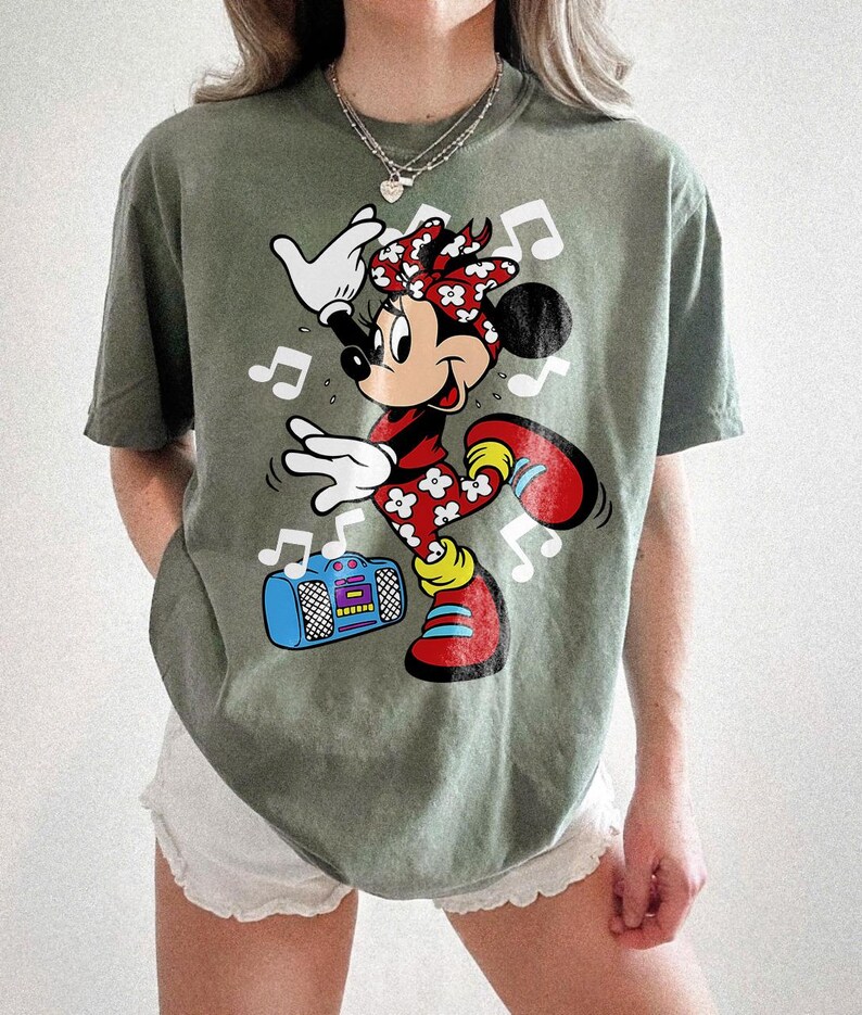 Pu&ograve; includere: T-shirt verde oliva con una grafica di Minnie Mouse che balla con note musicali e un boombox. Minnie Mouse indossa un vestito floreale rosso e bianco, scarpe rosse e un fiocco abbinato. Il boombox &egrave; blu con dettagli viola e gialli.