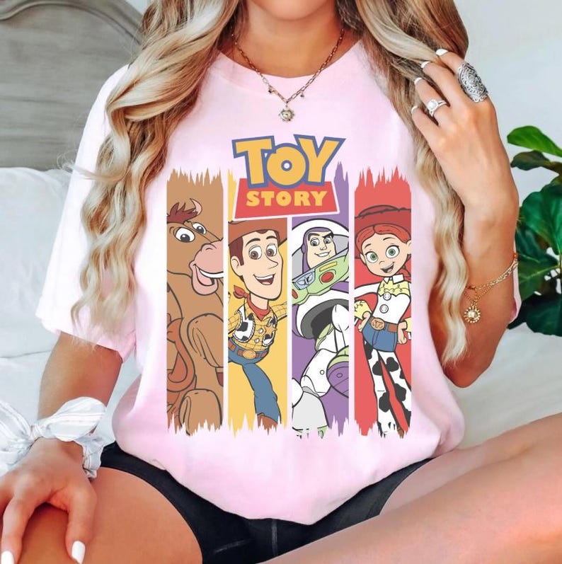 May include: Pink t-shirt featuring the words "TOY STORY" above cartoon images of characters. The characters include a brown horse, Woody, Buzz Lightyear, and Jessie, each in a vertical colored stripe.