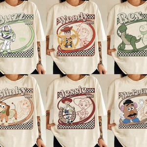 May include: Cream-colored t-shirts featuring various Toy Story characters: Buzz Lightyear, Woody, Rex, Slinky Dog, Jessie, and Mr. Potato Head. Each design includes the character's name and "1995" in a retro-style frame with a checkered border.