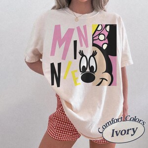 Op de afbeelding: Ivoor kleurig T-shirt met een afbeelding van Minnie Mouse en het woord "MINNIE" in roze, zwarte en gele blokletters. Het shirt is van het merk Comfort Colors.