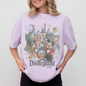 Puede incluir: Camiseta lila claro con un gr&aacute;fico de Disneyland que presenta a Mickey Mouse, Minnie Mouse, Donald Duck, Goofy y Pluto. El gr&aacute;fico incluye un castillo, fuegos artificiales y una noria. El texto "Disneyland Est. 1955" est&aacute; impreso debajo.