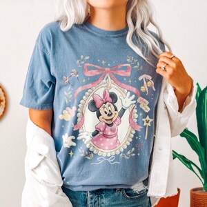 May include: A blue t-shirt featuring a graphic of Minnie Mouse inside an ornate, pearl-bordered oval frame. The design includes a large pink bow above the frame, small star accents, and floral elements. Minnie is wearing a pink polka-dot dress.