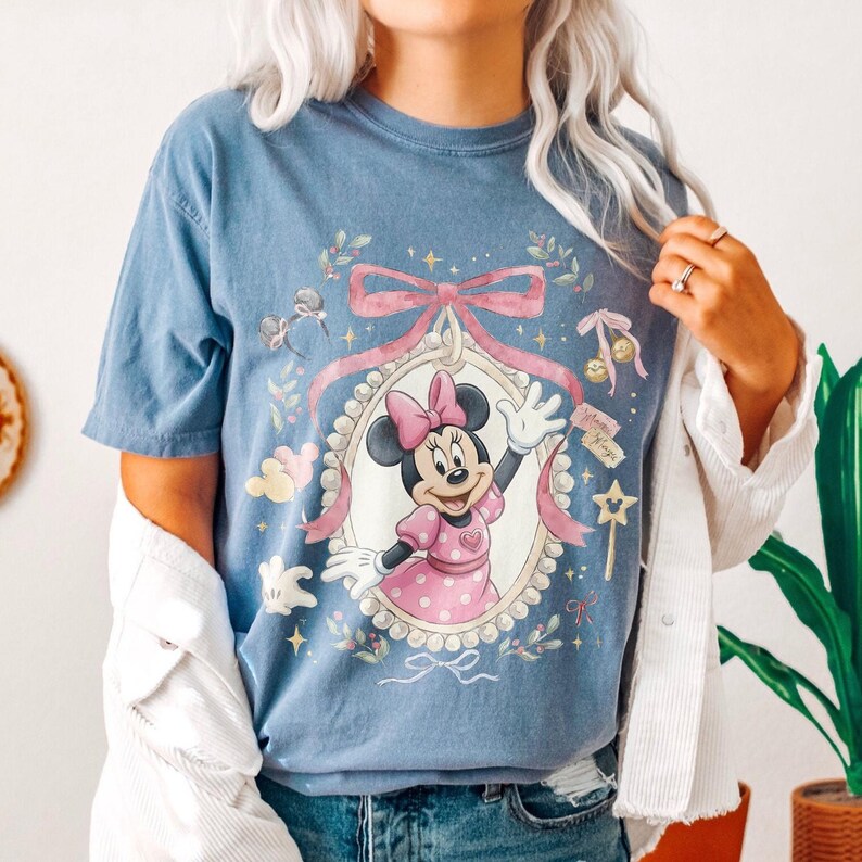 May include: A blue t-shirt featuring a graphic of Minnie Mouse inside an ornate, pearl-bordered oval frame. The design includes a large pink bow above the frame, small star accents, and floral elements. Minnie is wearing a pink polka-dot dress.