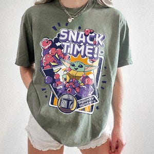 May include: An olive green short-sleeve t-shirt featuring a graphic print with the text "SNACK TIME!" and "Bounty Hunters". The design includes a small green creature with large ears eating purple and red snacks, surrounded by a stylized helmet icon.