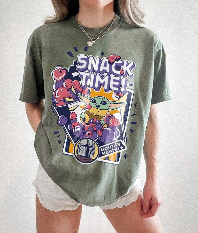 May include: An olive green short-sleeve t-shirt featuring a graphic print with the text "SNACK TIME!" and "Bounty Hunters". The design includes a small green creature with large ears eating purple and red snacks, surrounded by a stylized helmet icon.