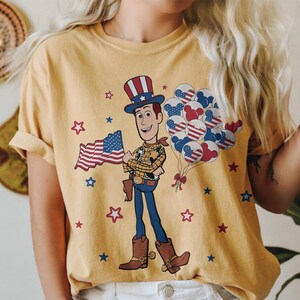May include: Mustard yellow t-shirt featuring a cartoon Woody character holding an American flag and balloons. The balloons are designed with red, white, and blue patterns. Red and blue stars are scattered around the graphic.