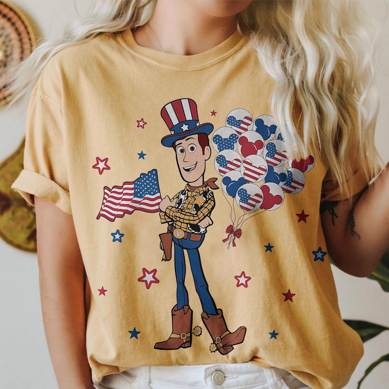 May include: Mustard yellow t-shirt featuring a cartoon Woody character holding an American flag and balloons. The balloons are designed with red, white, and blue patterns. Red and blue stars are scattered around the graphic.