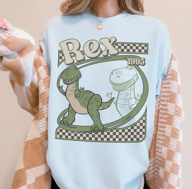 May include: Light blue sweatshirt featuring a green cartoon dinosaur named Rex, with the text "Rex 1995" in a retro font. The design includes a checkered pattern and a second outline of Rex. A fun, playful design.