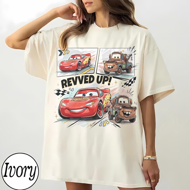 May include: An ivory t-shirt featuring a graphic print of Lightning McQueen and Mater from the movie Cars. The design includes two smaller panels at the top and a larger central illustration with the text REVVED UP! and checkered flag accents.