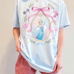May include: A light blue t-shirt featuring a central oval frame decorated with pearls and a pink bow. Inside the frame is an illustration of a blonde character in a blue gown. Surrounding the frame are small icons including mouse ears, cherries, balloons, and a star wand.