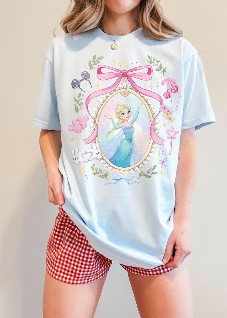 May include: A light blue t-shirt featuring a central oval frame decorated with pearls and a pink bow. Inside the frame is an illustration of a blonde character in a blue gown. Surrounding the frame are small icons including mouse ears, cherries, balloons, and a star wand.