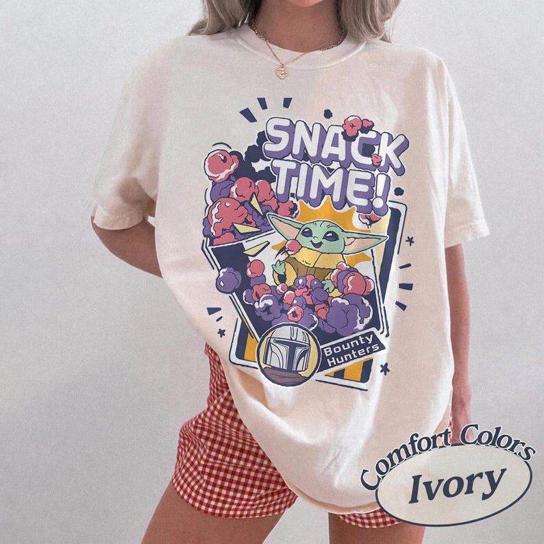 May include: An ivory short-sleeve t-shirt featuring a graphic print with the text SNACK TIME! and Bounty Hunters. The design includes a green character eating a snack surrounded by purple and pink clouds, with a helmet icon below the main graphic.