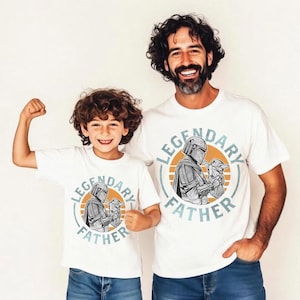 K&ouml;nnte beinhalten: Zwei wei&szlig;e T-Shirts mit dem Text "LEGENDARY FATHER" und einer Grafik eines Charakters, der ein Baby h&auml;lt. Die Shirts werden von einem Mann und einem Kind getragen. Die Grafik befindet sich in einem Kreis mit orangefarbenem Hintergrund.