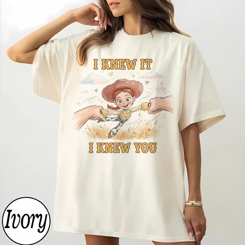 May include: An ivory t-shirt featuring a graphic print of a cartoon cowgirl character with red hair and a brown hat, being held by two hands. The text "I KNEW IT" appears above the character and "I KNEW YOU" appears below in a yellow font.