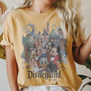 Puede incluir: Camiseta amarilla mostaza con un gr&aacute;fico de Disneyland de estilo vintage. El dise&ntilde;o incluye a Mickey Mouse, Minnie Mouse, Donald Duck y otros personajes de Disney frente a un castillo y fuegos artificiales. El texto "Disneyland Est.1955" est&aacute; impreso debajo.