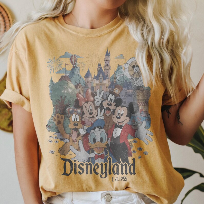 Puede incluir: Camiseta amarilla mostaza con un gr&aacute;fico de Disneyland de estilo vintage. El dise&ntilde;o incluye a Mickey Mouse, Minnie Mouse, Donald Duck y otros personajes de Disney frente a un castillo y fuegos artificiales. El texto "Disneyland Est.1955" est&aacute; impreso debajo.