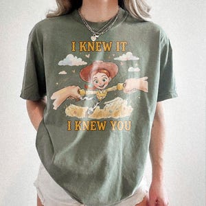 May include: An olive green short-sleeve t-shirt featuring a graphic of a cowgirl character with red hair and a brown hat. The text I KNEW IT and I KNEW YOU is printed in yellow above and below the character, who is reaching out to two hands.