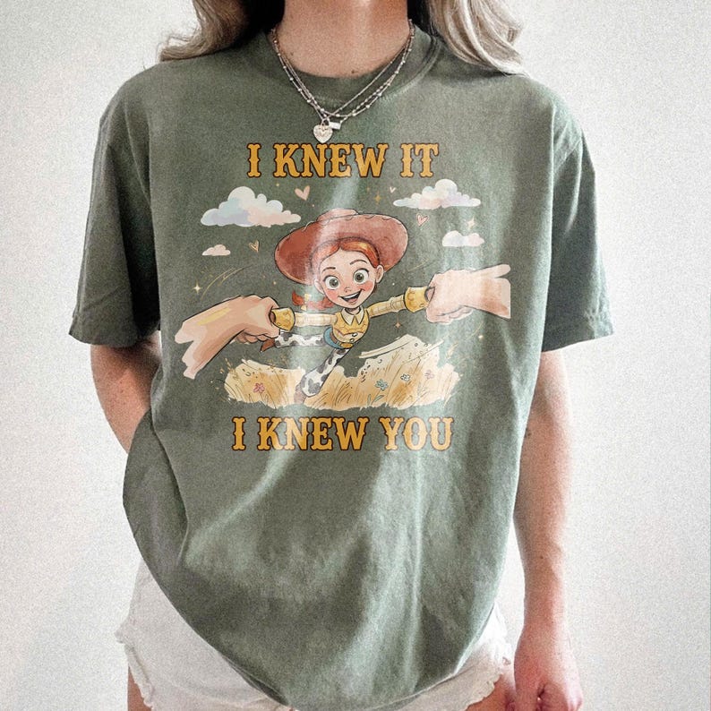 May include: An olive green short-sleeve t-shirt featuring a graphic of a cowgirl character with red hair and a brown hat. The text I KNEW IT and I KNEW YOU is printed in yellow above and below the character, who is reaching out to two hands.