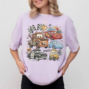 May include: A light purple short-sleeve t-shirt featuring a colorful graphic print of various cartoon cars, including a brown tow truck, a red race car, a blue sports car, a black police car, and a yellow forklift, with the text GO! and SPEED!
