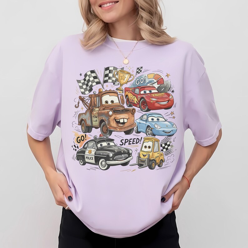 May include: A light purple short-sleeve t-shirt featuring a colorful graphic print of various cartoon cars, including a brown tow truck, a red race car, a blue sports car, a black police car, and a yellow forklift, with the text GO! and SPEED!