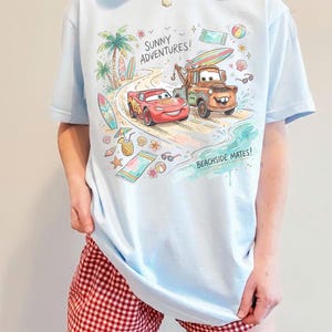 May include: A light blue t-shirt featuring a colorful graphic of Lightning McQueen and Mater from the movie Cars on a beach. The design includes palm trees, surfboards, and the text SUNNY ADVENTURES! and BEACHSIDE MATES! in a playful font.