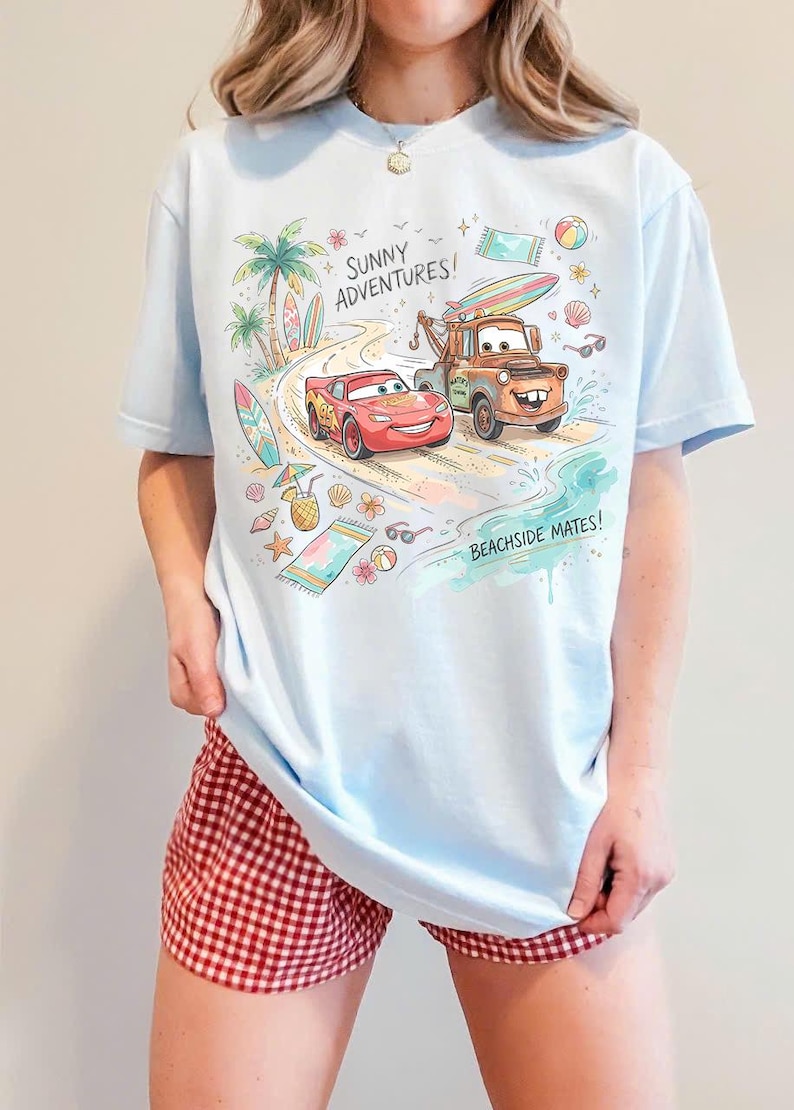 May include: A light blue t-shirt featuring a colorful graphic of Lightning McQueen and Mater from the movie Cars on a beach. The design includes palm trees, surfboards, and the text SUNNY ADVENTURES! and BEACHSIDE MATES! in a playful font.