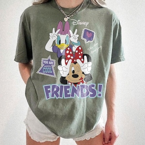 Może przedstawiać: Oliwkowa koszulka z Myszką Minnie i Daisy Duck z Disneya. Myszka Minnie ma czerwoną kokardę, a Daisy Duck fioletową kokardę. Koszulka ma napis "FRIENDS!" w fioletowych literach i tekst "You Are Cool".