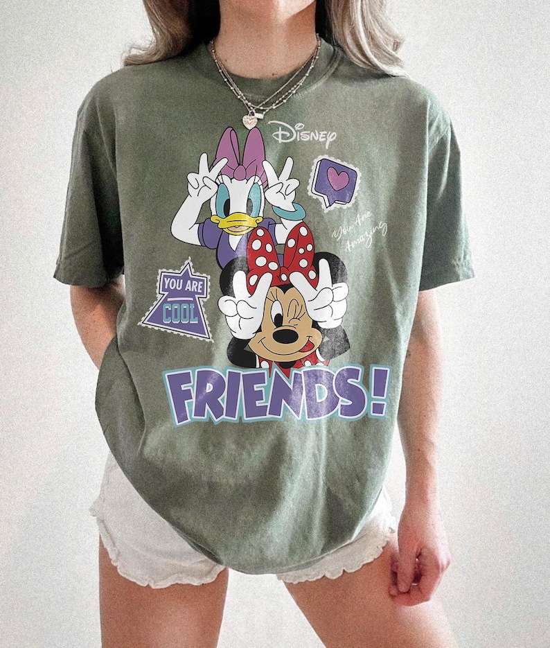 Może przedstawiać: Oliwkowa koszulka z Myszką Minnie i Daisy Duck z Disneya. Myszka Minnie ma czerwoną kokardę, a Daisy Duck fioletową kokardę. Koszulka ma napis "FRIENDS!" w fioletowych literach i tekst "You Are Cool".