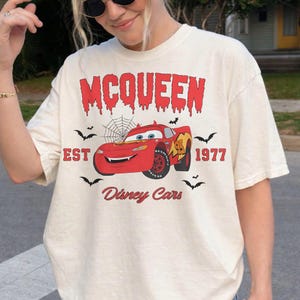 May include: Cream-colored t-shirt featuring a graphic of Lightning McQueen from Disney Cars. The design includes the word "McQueen" in red, the text "EST 1977", and several bats. The shirt is a relaxed fit.