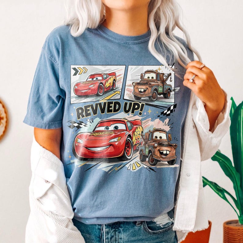 May include: A blue t-shirt featuring a graphic print of the characters Lightning McQueen and Mater from the movie Cars. The design includes the text REVVED UP! with comic-style panels showing the red race car and brown tow truck in action.