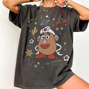 May include: Dark gray t-shirt featuring a cartoon Mrs. Potato Head design. The graphic includes a brown potato head with a white hat, red lips, and gold earrings. Additional elements include stars, a cactus, and the text "Mrs. Potato".