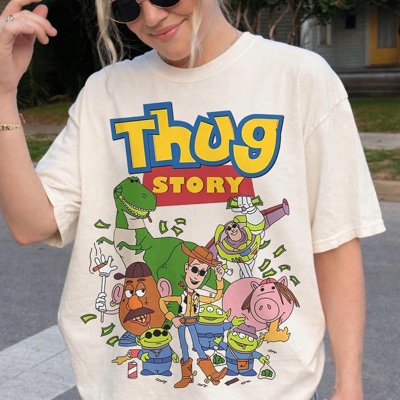 Pode incluir: Camiseta creme com um gr&aacute;fico de personagens de desenhos animados e o texto "Thug Story" em amarelo e azul. O design inclui um dinossauro verde, Woody, Buzz Lightyear e outros personagens com s&iacute;mbolos de dinheiro.