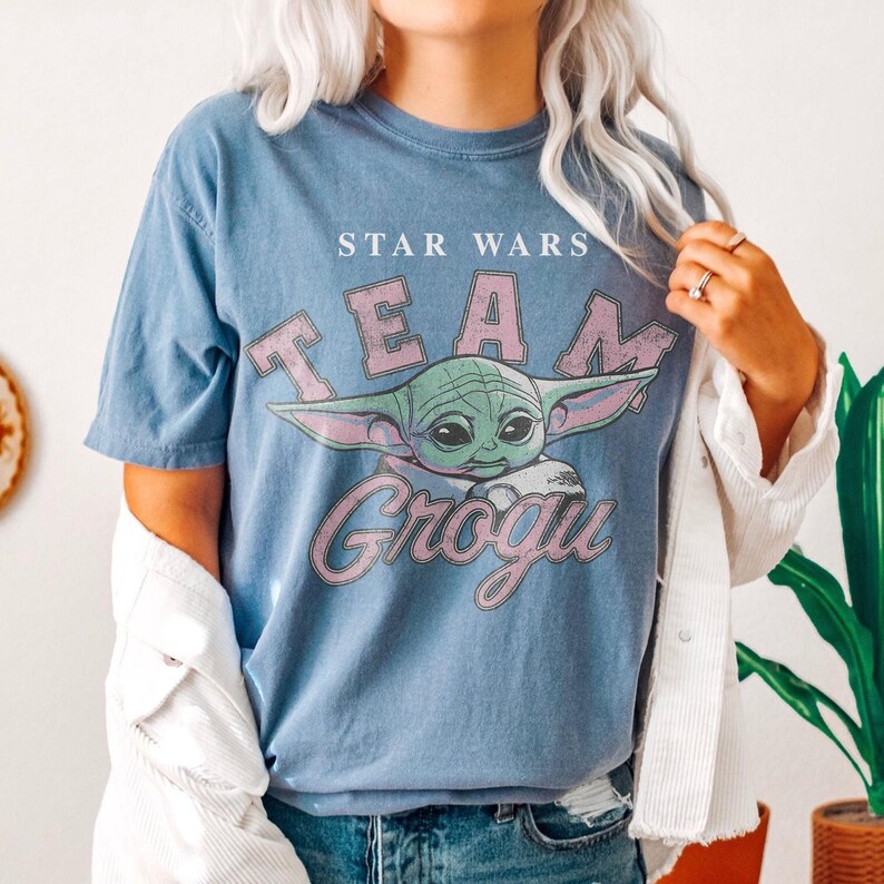 Puede incluir: Una camiseta azul de manga corta con un gr&aacute;fico del personaje Grogu de Star Wars. El texto dice STAR WARS TEAM GROGU en una fuente rosa desgastada. La camiseta tiene cuello redondo y un corte relajado, usada debajo de una chaqueta de pana blanca.