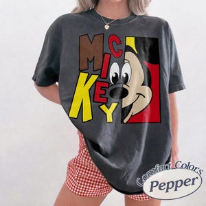 Op de afbeelding: Donkergrijs t-shirt met een Mickey Mouse-afbeelding en het woord "MICKEY" in bruin, rood en geel. Het shirt is van het merk Comfort Colors.