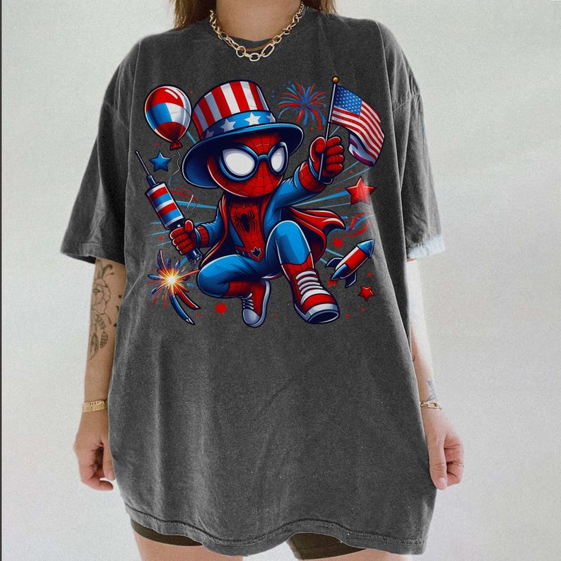 K&ouml;nnte beinhalten: Dunkelgraues T-Shirt mit einem patriotischen Spider-Man-Charakter, der eine amerikanische Flagge, Feuerwerk und eine Rakete h&auml;lt. Die Figur tr&auml;gt einen rot-wei&szlig;-blauen Hut und Anzug. Das Design umfasst Sterne und einen Ballon.