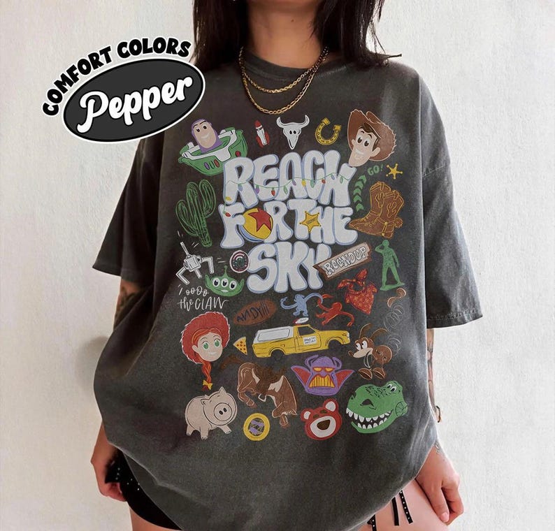 May include: A charcoal grey short-sleeve t-shirt featuring a graphic print with the text Reach For The Sky and various cartoon character illustrations including a cowboy, space ranger, green alien, cowgirl, pig, dinosaur, and a yellow truck.