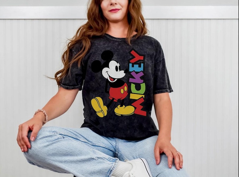 May include: Black acid wash t-shirt featuring a colorful graphic of Mickey Mouse with the word "MICKEY" in rainbow colors. The iconic cartoon character is depicted in his classic pose, with red shorts and yellow shoes.