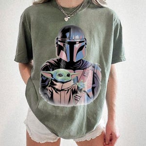 May include: Olive green t-shirt featuring a graphic of a Mandalorian helmet and Grogu. The illustration has blue, pink, and black accents. The t-shirt is a relaxed fit.