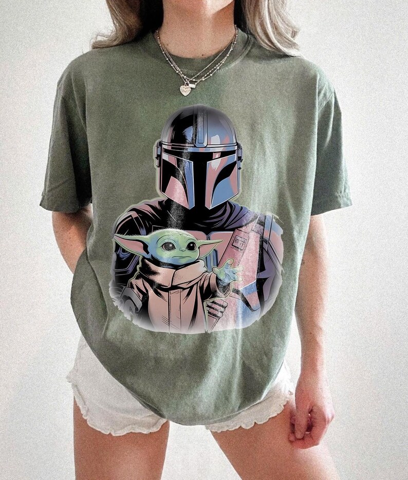 May include: Olive green t-shirt featuring a graphic of a Mandalorian helmet and Grogu. The illustration has blue, pink, and black accents. The t-shirt is a relaxed fit.