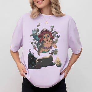 May include: A light purple short-sleeved t-shirt featuring a graphic print of the character Ariel from The Little Mermaid, surrounded by other characters including King Triton, Flounder, and Sebastian, with an underwater castle scene below.