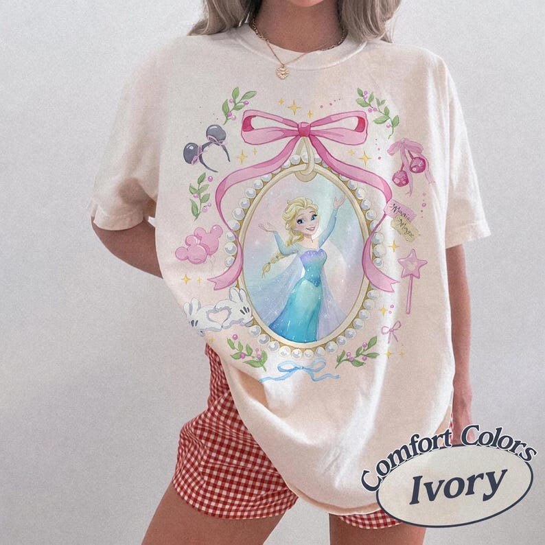 May include: An ivory short-sleeve t-shirt featuring a central graphic of a blonde character in a blue gown inside a pearl-bordered oval frame. The design includes a large pink bow, cherry icons, mouse ears, and star motifs in pastel colors.
