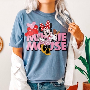 May include: A blue t-shirt featuring a graphic of Minnie Mouse holding a chocolate bar and balloons. The text "MINNIE MOUSE" is in pink. Minnie Mouse is wearing a polka dot dress and a red bow.