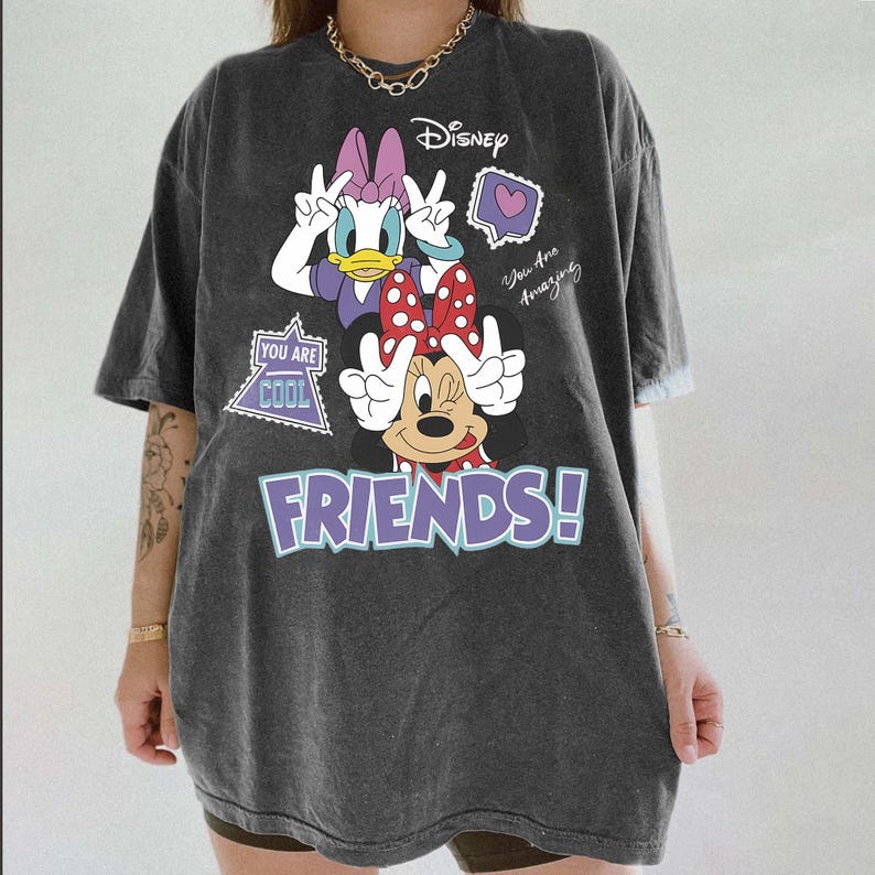 Może przedstawiać: Ciemnoszara koszulka z Daisy Duck i Minnie Mouse pokazującymi znak pokoju. Koszulka marki Disney ma fioletowy napis "FRIENDS!" wraz z frazami "You Are Cool" i "You Are Amazing."