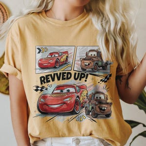 May include: A mustard yellow t-shirt featuring a graphic print of Lightning McQueen and Mater from the movie Cars. The design includes comic-style panels, checkered flags, and the text REVVED UP! in bold black letters across the center.