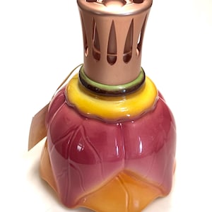 May include: A ceramic fragrance lamp shaped like a flower with pink and orange petals, a yellow center, and a metallic rose gold-colored vented cap on top. A small paper tag is attached to the neck of the lamp, which features a glossy finish.